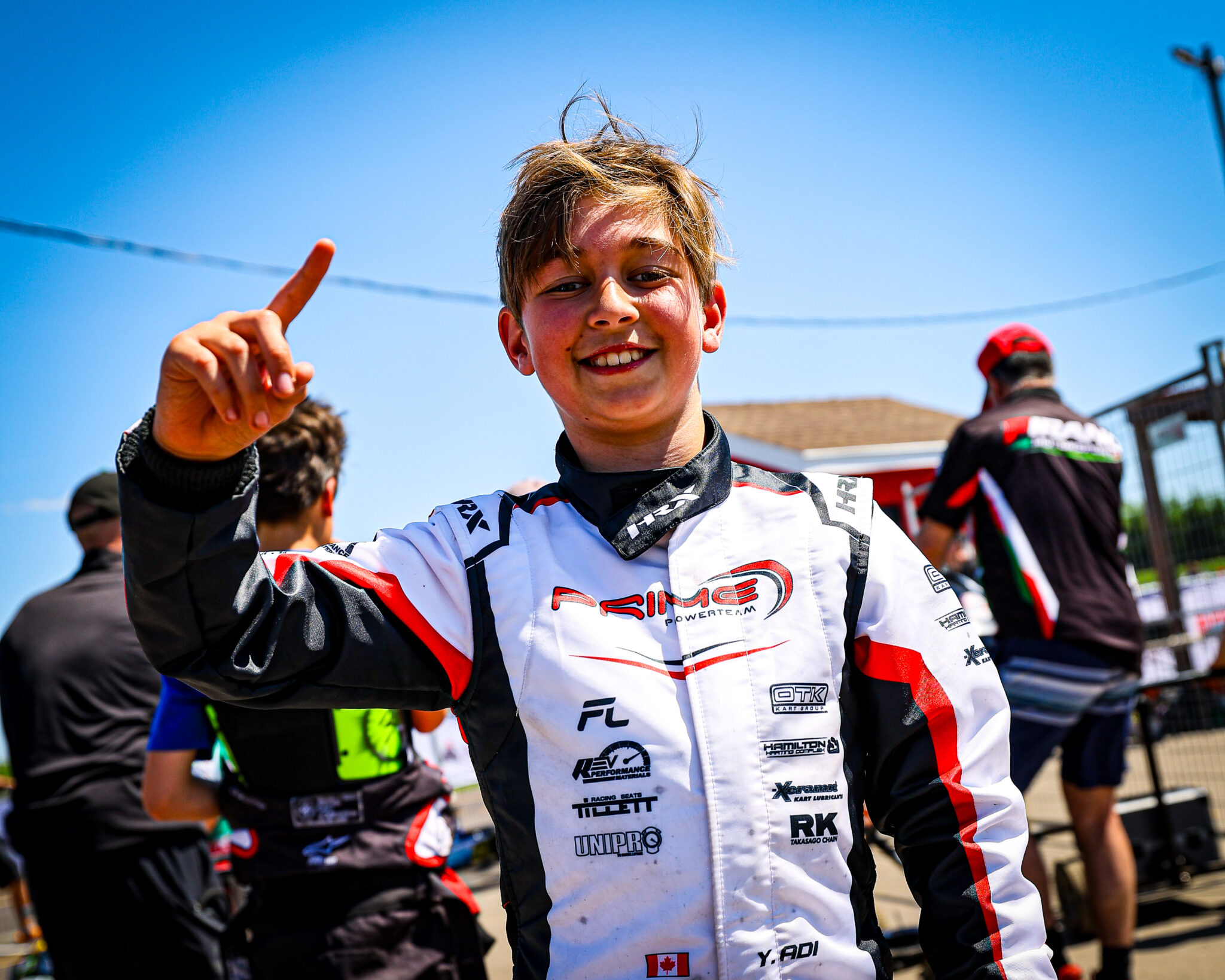 Smiling young racer in a white, red, and black racing suit giving a thumbs up outdoors against a blue sky.