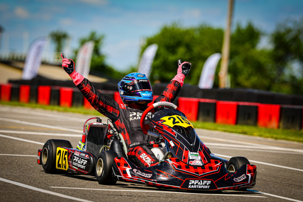 Go-kart racer in red and black suit with blue helmet raises arms in victory on a race track, kart number 215 visible on the front panel.