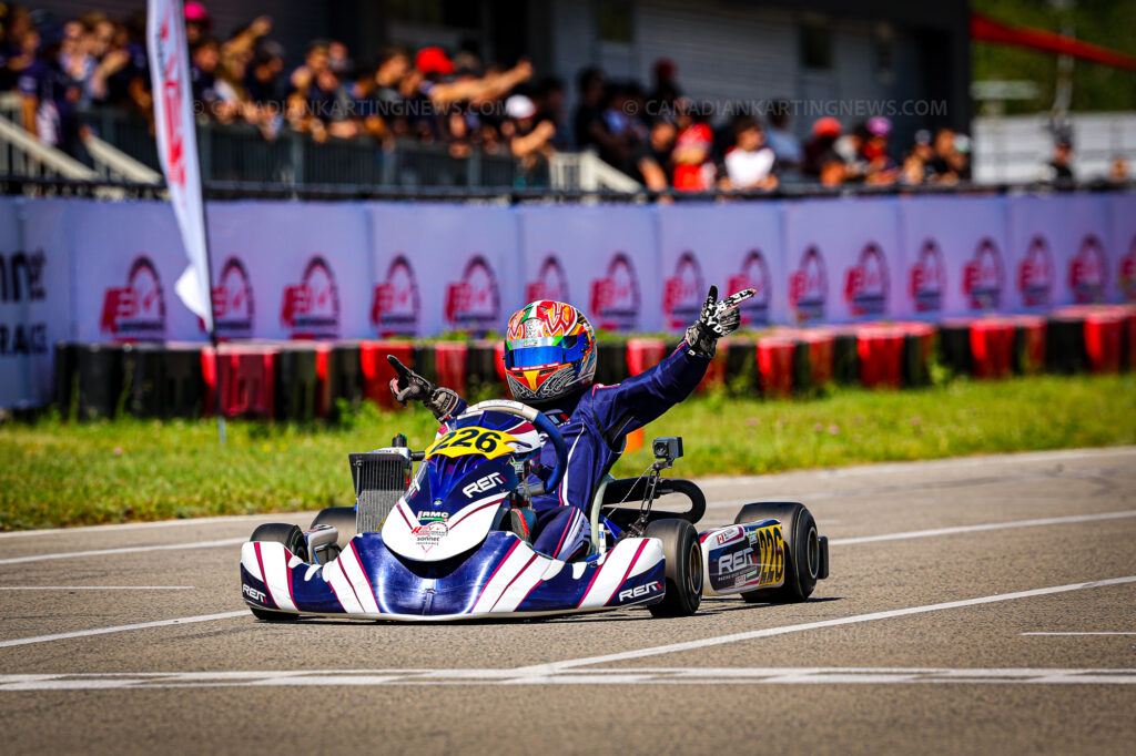 Go-kart racer in a blue suit and colorful helmet raises an arm in celebration as spectators cheer in the blurred background.
