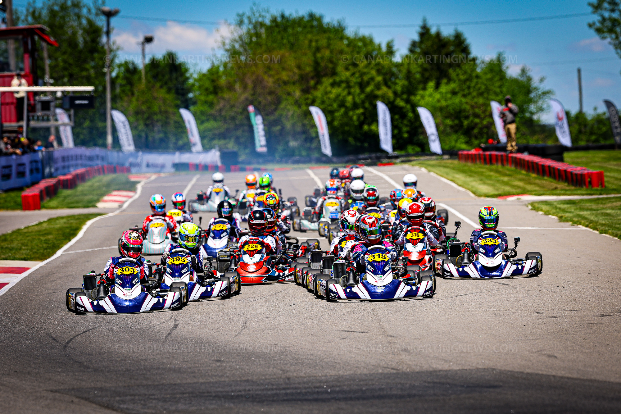 Group of go-kart racers at the start of a race, colorful helmets and numbered karts on a sunny track.",